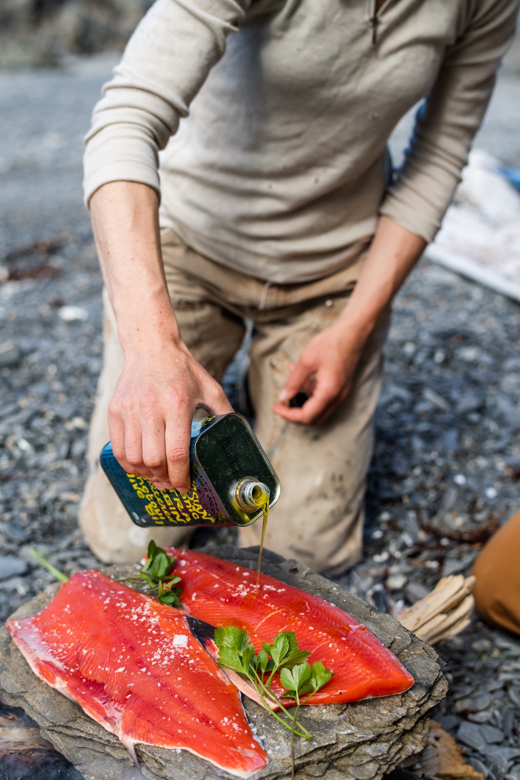 Kneeling on a rocky beach pouring olive oil from a tin over two salted sockeye fillets on a stone slab, photographed in Alaska