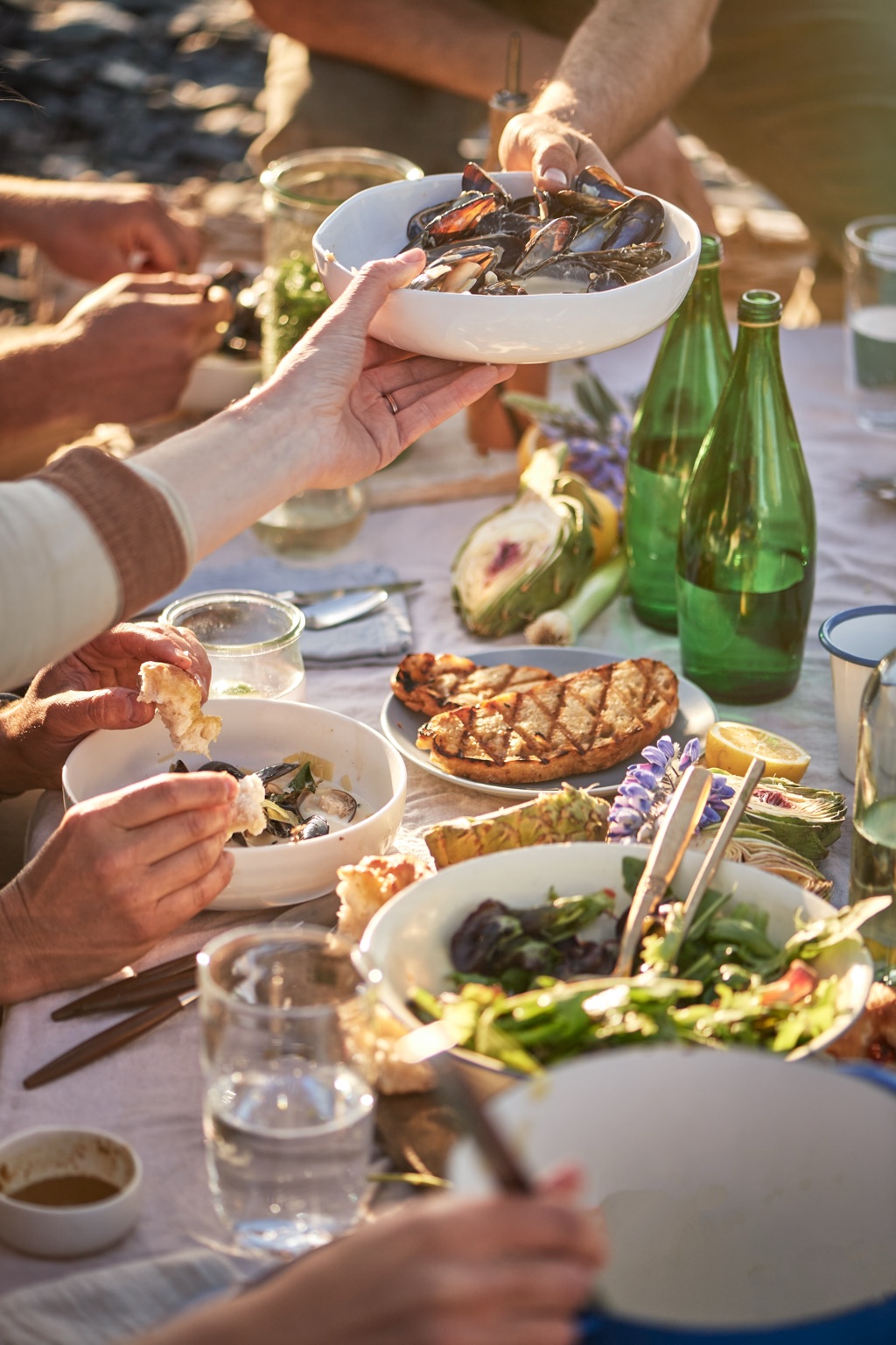 Passing a bowl of mussels across a sunlit beach dinner table with green glass bottles and grilled bread on the Alaska coast