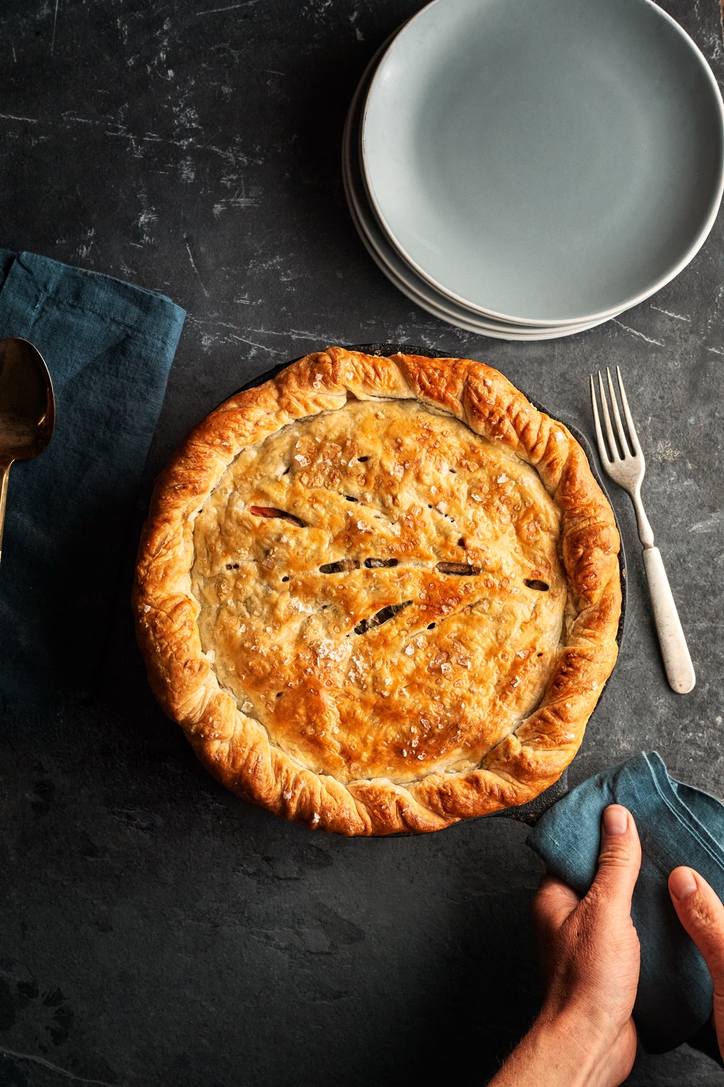 Golden flaky pot pie in a cast iron skillet with sea salt on the crust, a hand gripping the handle with a linen napkin