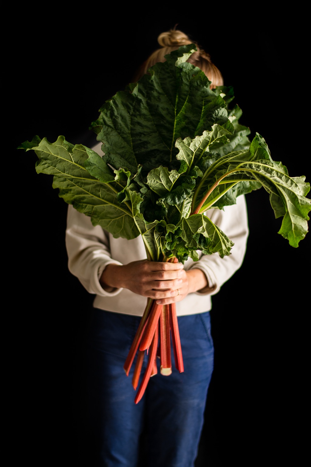 Woman holding a giant leafy bunch of rhubarb in front of her face against a black background