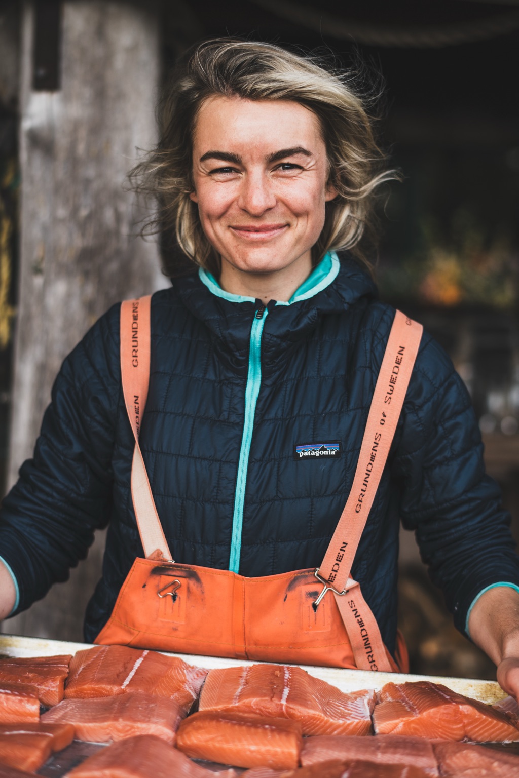 Portrait of a fisherwoman in a navy Patagonia puffy and orange Grundens bibs behind a tray of fresh salmon fillets, photographed in Alaska, photographed by Dawn Heumann