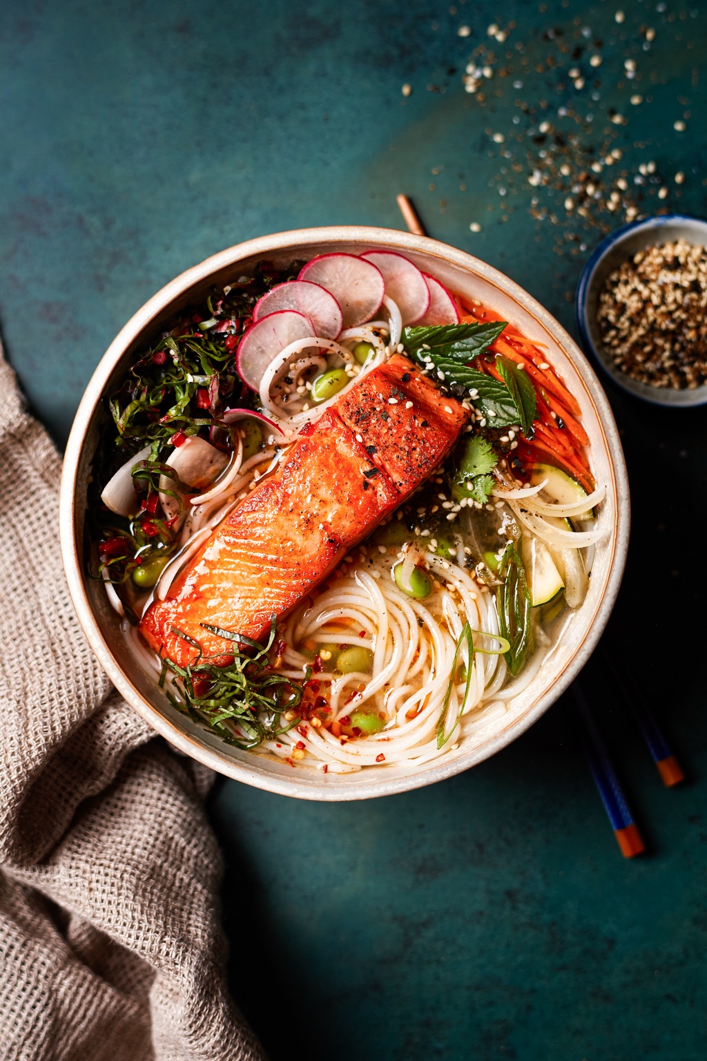 Overhead bowl of seared salmon over rice noodles with radishes, carrots, edamame, mint, and sesame on a teal backdrop, photographed in Alaska