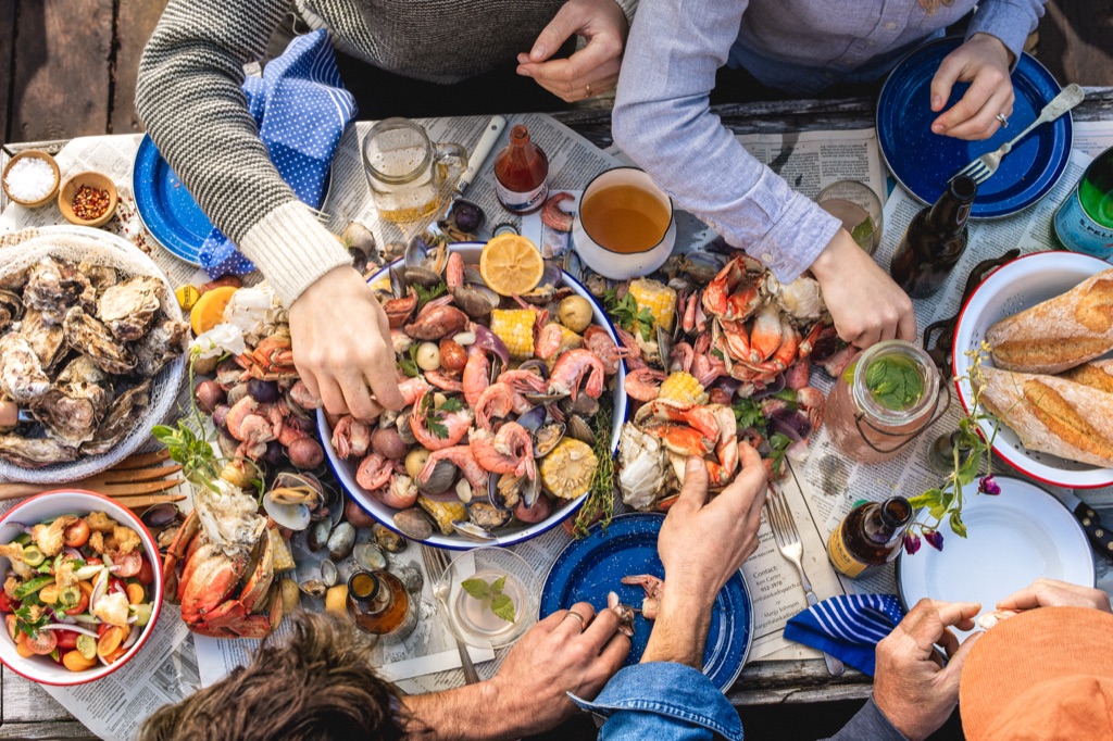 Hands reaching into a shared platter of Dungeness crab, spot prawns, clams, and corn at an outdoor seafood boil, photographed in Alaska