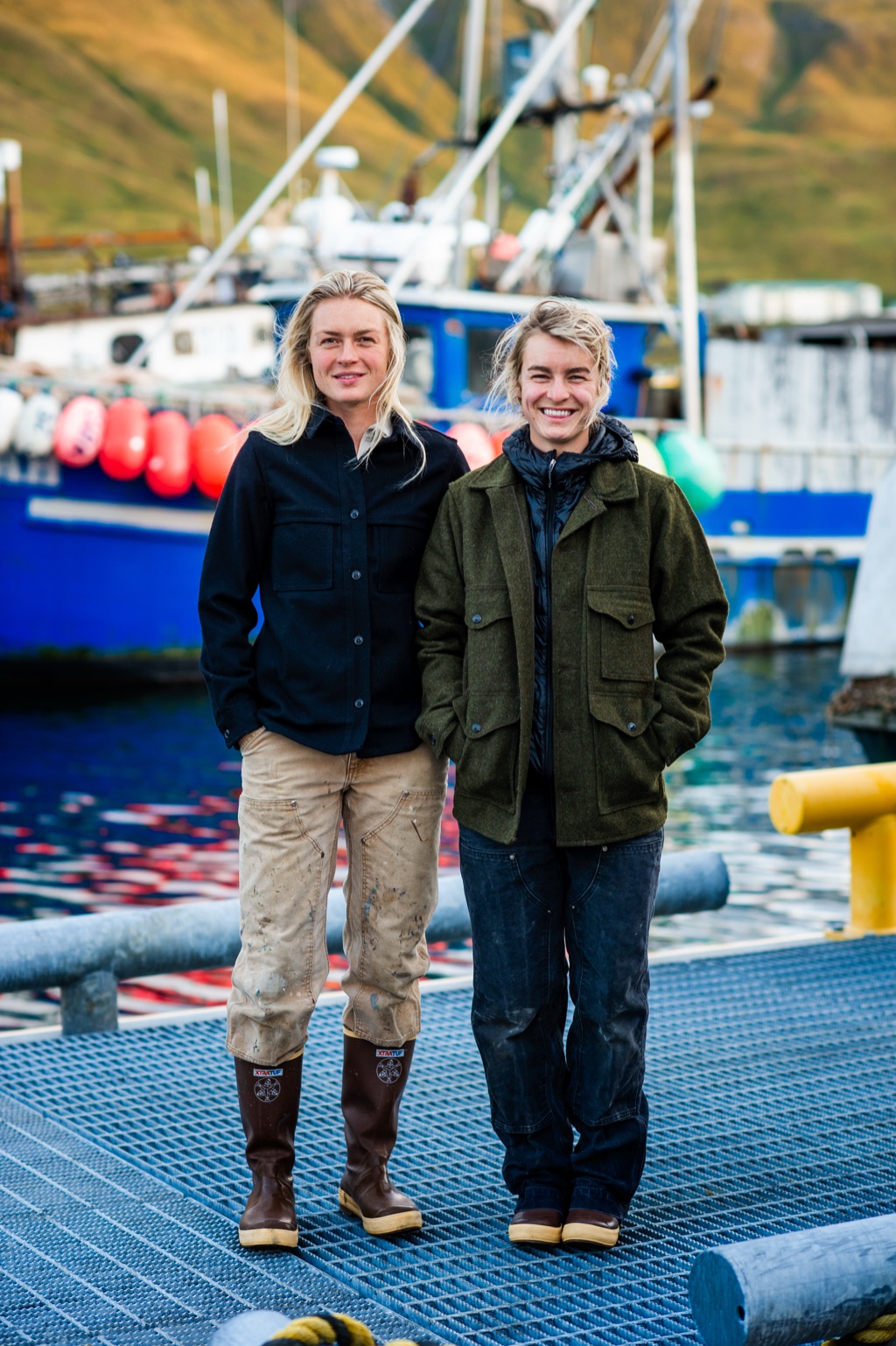Two fisherwomen in wool jackets and XtraTuf boots standing on a dock grate before a blue fishing boat with buoys in Alaska