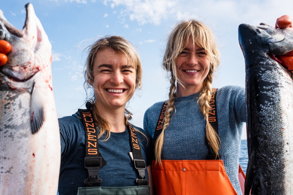 Selfie-style shot of two women in Grundens bibs each holding up a salmon against a blue sky, photographed in Alaska