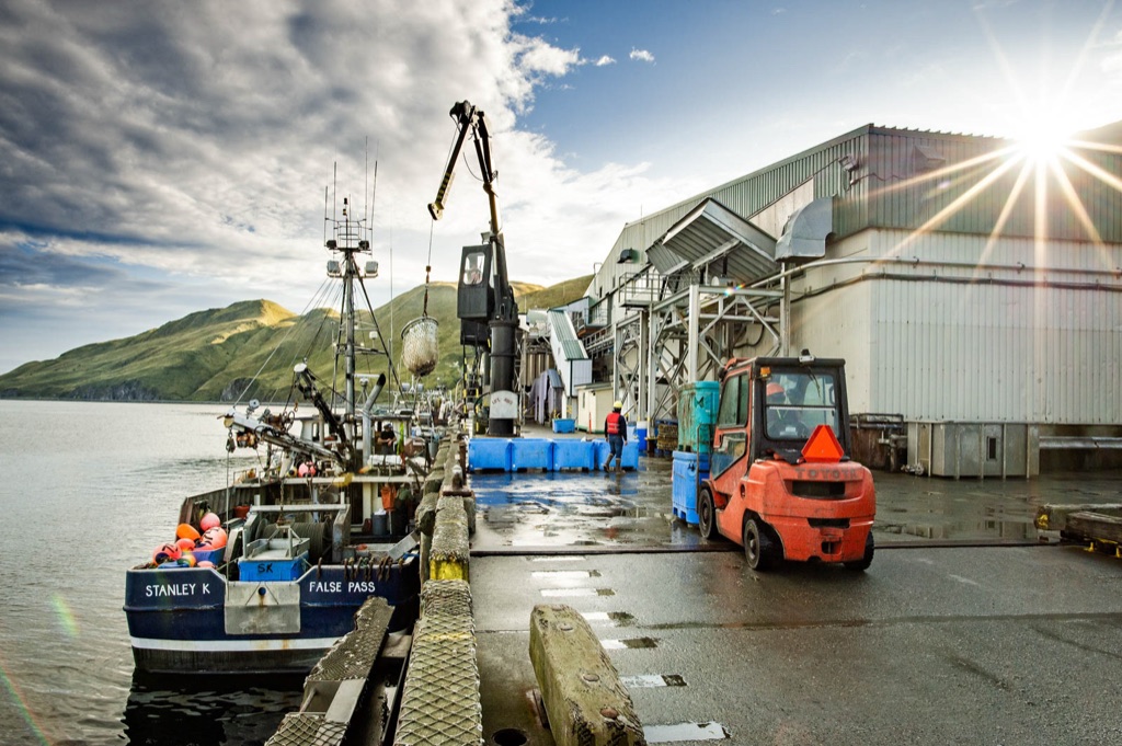 Fishing boat Stanley K of False Pass tied up at a cannery dock under a sunburst, crane and forklift on the pier in Alaska