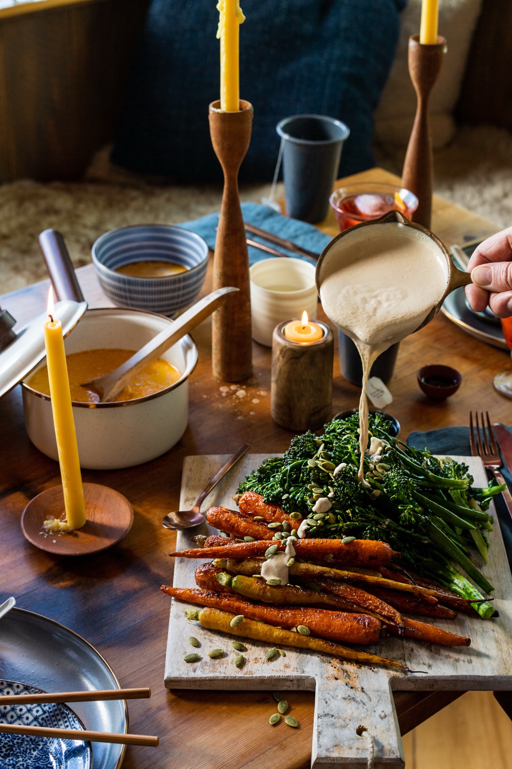 Pouring tahini sauce from a small copper pot over roasted carrots and broccolini on a candlelit table in Alaska
