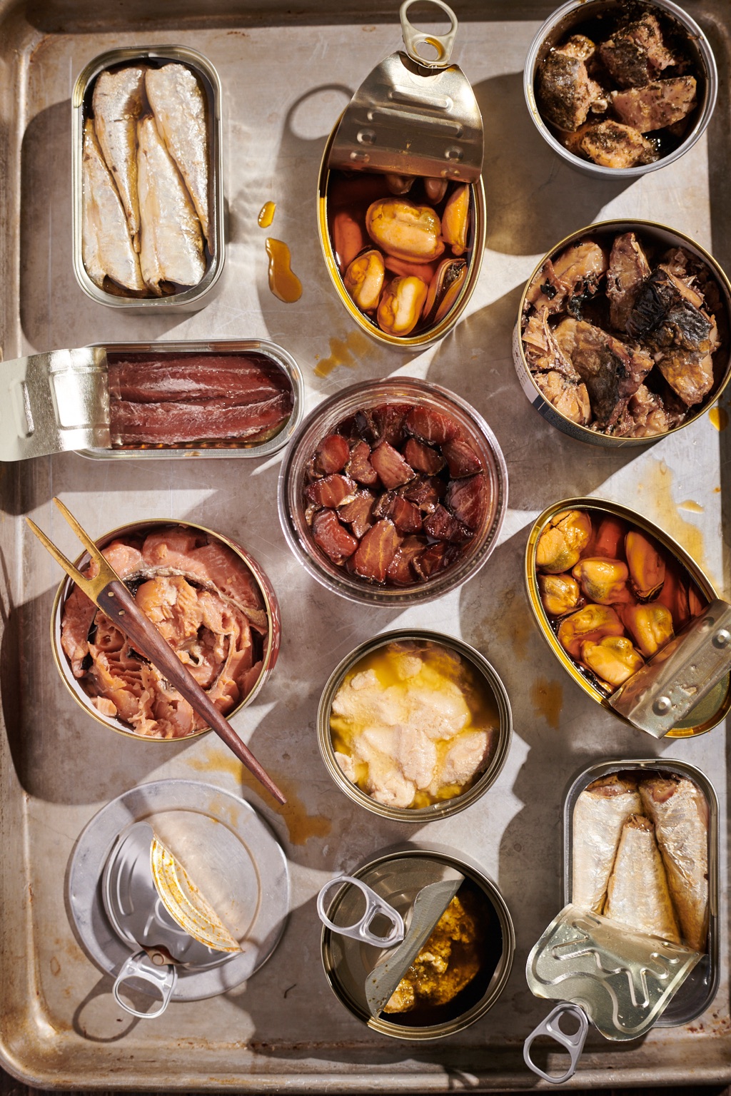 Opened tins of smoked salmon, sardines, mussels, mackerel, and anchovies arranged on a vintage sheet pan, photographed in Alaska