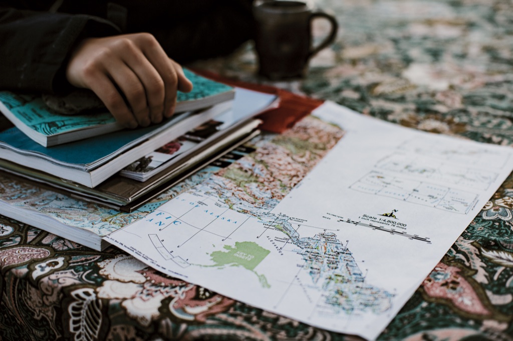 Hand resting on a stack of journals beside a map of Alaska and a mug of coffee
