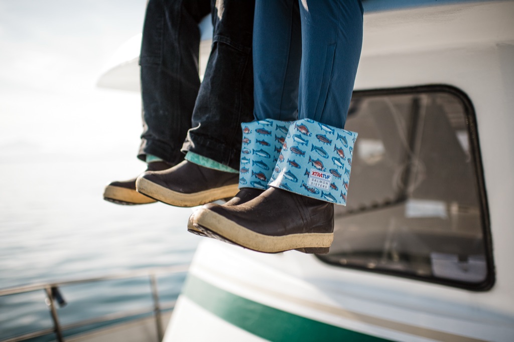 Legs dangling off a boat cabin in XtraTuf Salmon Sisters boots, one cuff rolled to show the fish print lining
