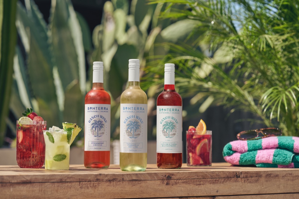 Three Ranch Wine bottles and garnished wine cocktails on a wood table with a striped towel and tropical plants