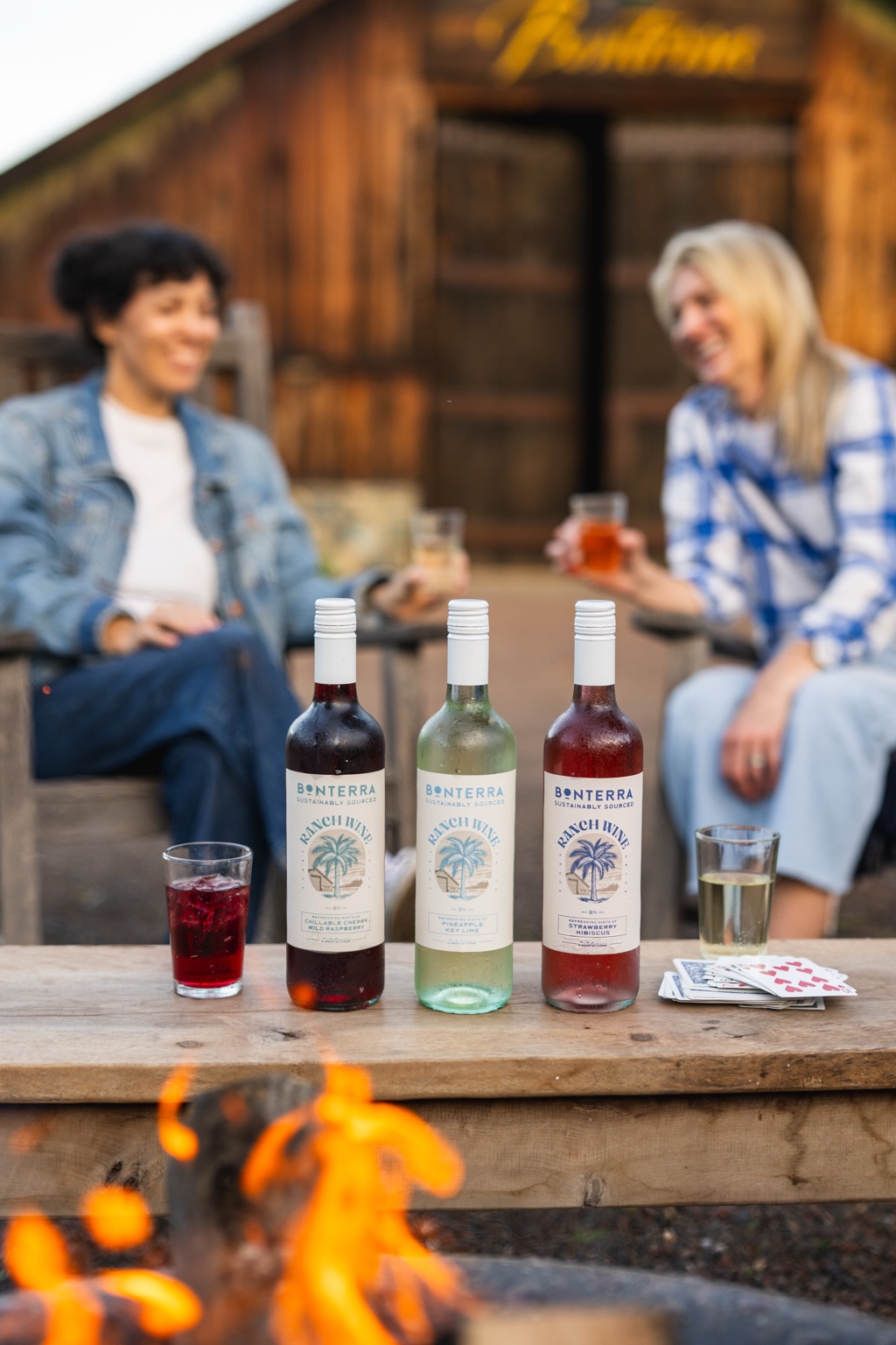 Two friends laughing by a campfire behind three Bonterra Ranch Wine bottles at the ranch, with playing cards and glasses on the table