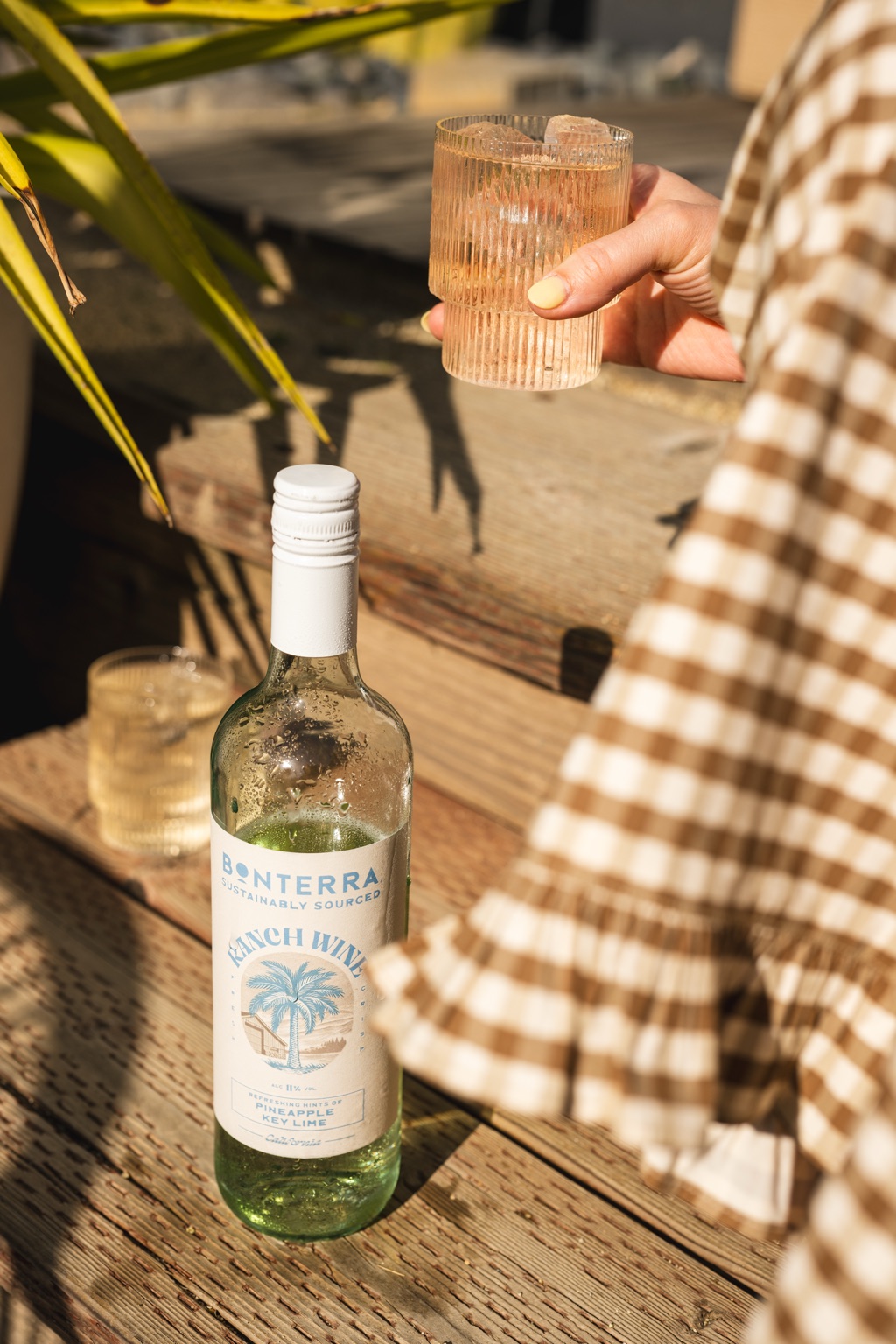 Woman in a gingham dress lifting an iced glass of Ranch Wine above a Pineapple Key Lime bottle on a weathered deck
