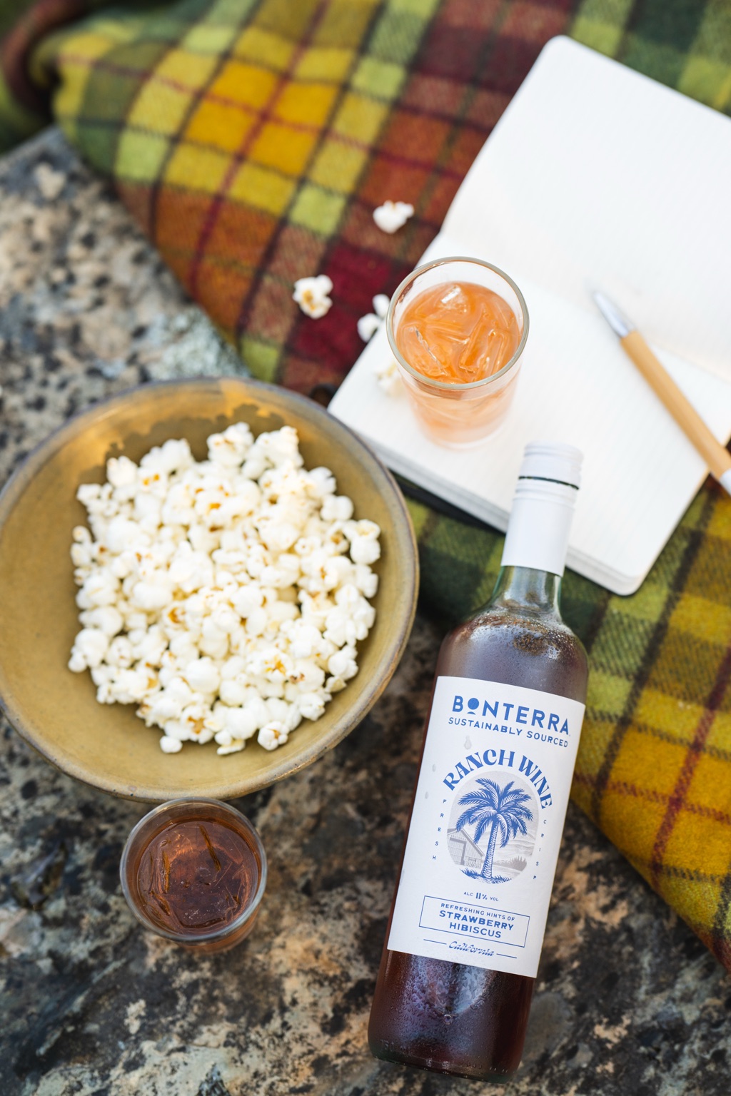 Overhead view of a Ranch Wine Strawberry Hibiscus bottle on a plaid picnic blanket with popcorn, a notebook, and iced glasses