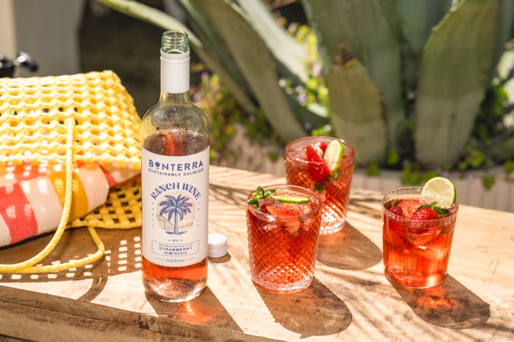 Opened bottle of Ranch Wine Strawberry Hibiscus with three textured glasses, a yellow woven bag, and a striped towel