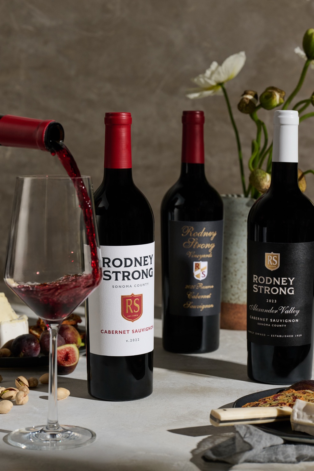 Red wine pouring into a glass beside Rodney Strong cabernet bottles, figs, and pistachios