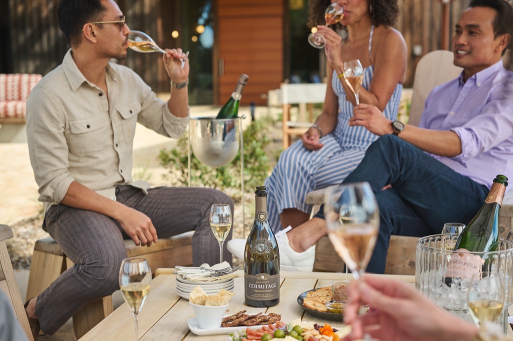 Friends sipping sparkling wine around a low table with L'Ermitage Brut, charcuterie, and an ice bucket