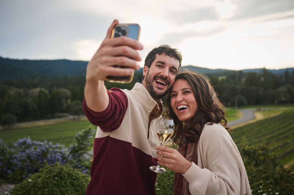 Laughing couple taking a selfie with a glass of sparkling wine above the vineyard at dusk