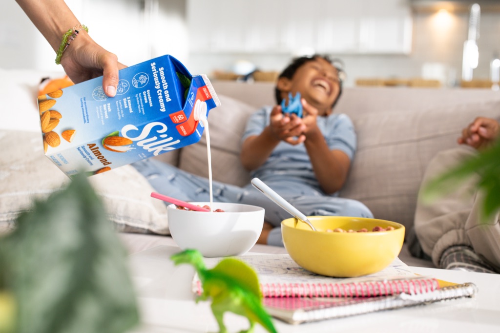 Silk almondmilk poured into a cereal bowl while a boy laughs with his dinosaur toys