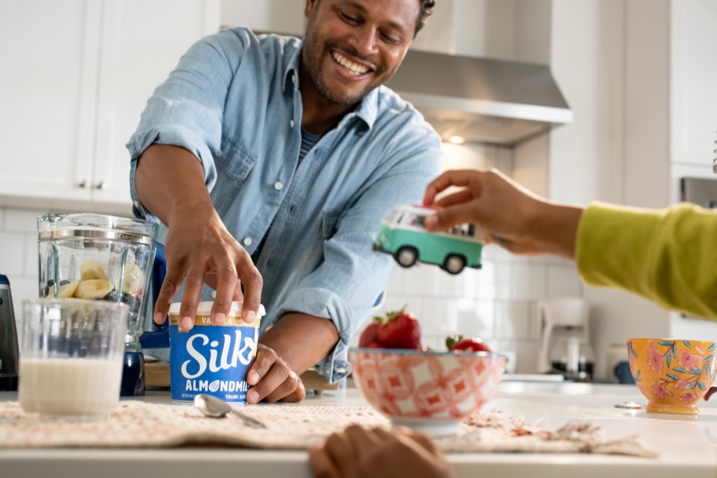 Dad grabbing Silk almondmilk yogurt at the blender while a kid trades him a toy bus