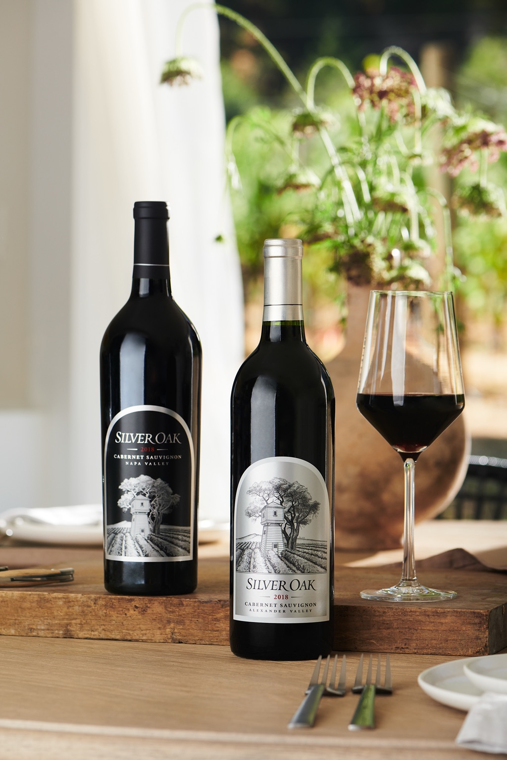 Silver Oak Napa Valley and Alexander Valley Cabernet Sauvignon bottles with a glass of red wine on a sunlit table, photographed by Dawn Heumann