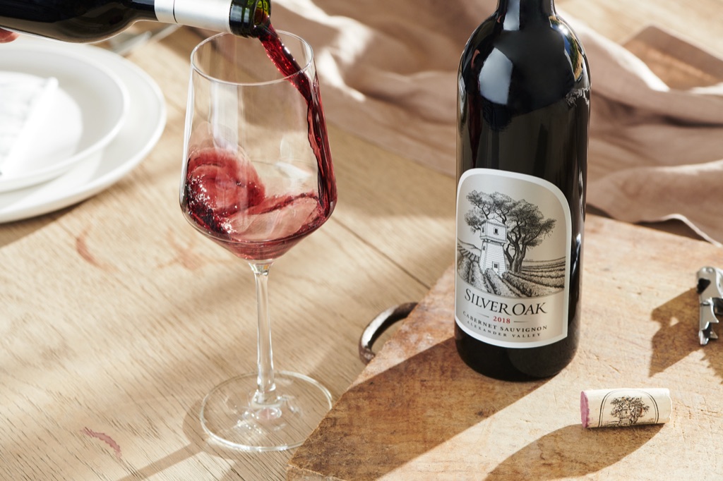 Cabernet swirling into a glass next to a Silver Oak Alexander Valley bottle and illustrated cork on a wood board
