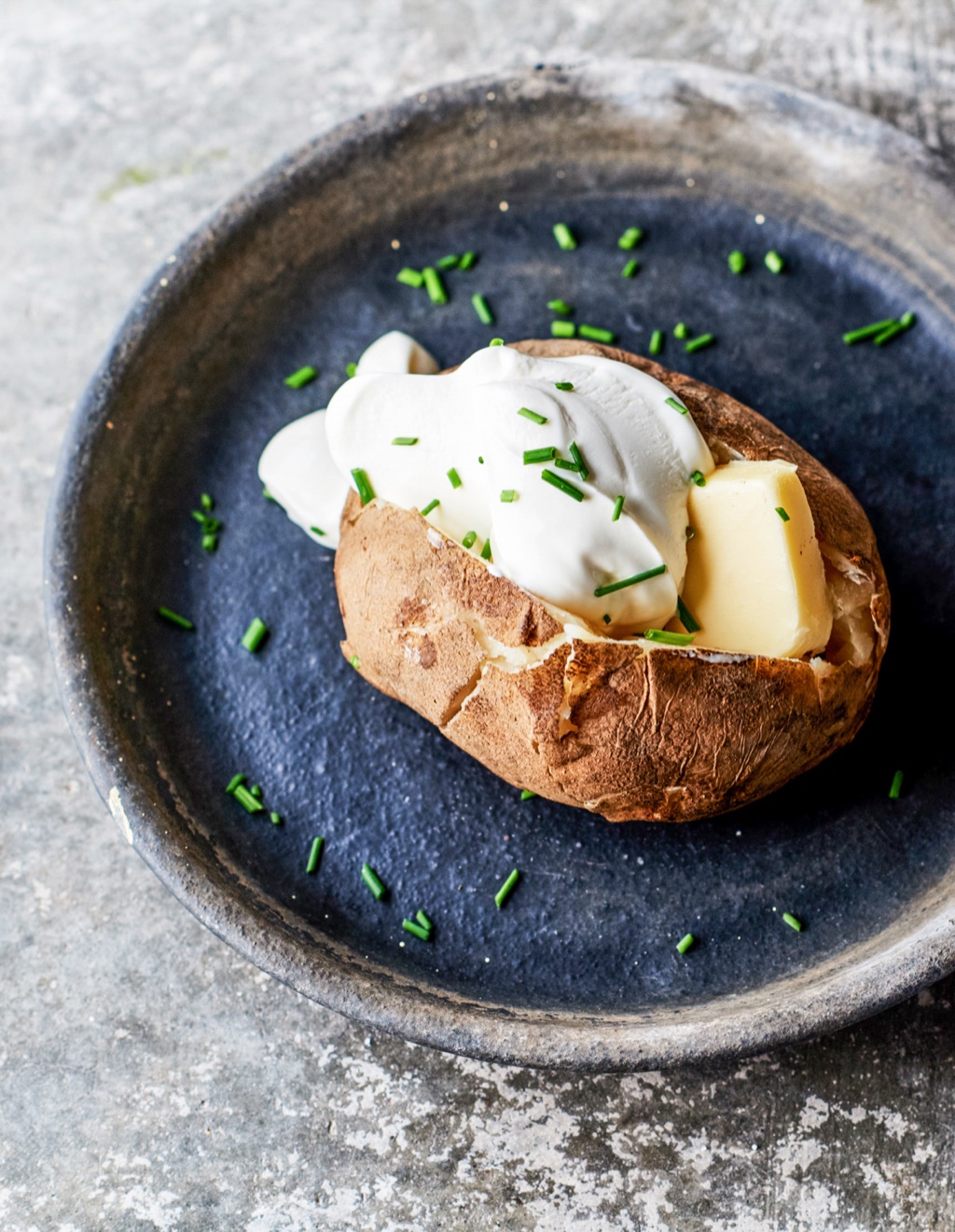 Baked potato loaded with sour cream, butter, and chives on a dark blue plate