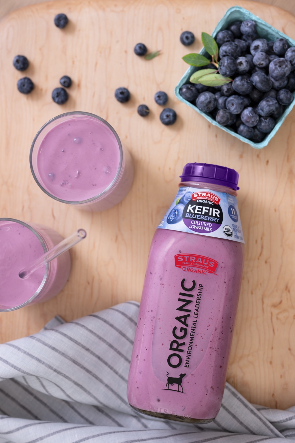 Overhead of a Straus Blueberry Kefir bottle with smoothie glasses and a basket of blueberries