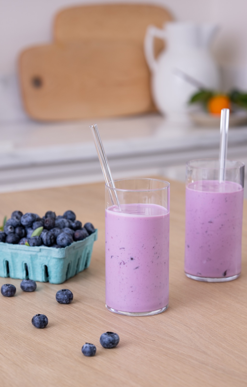 Two blueberry kefir smoothies with glass straws next to a carton of blueberries