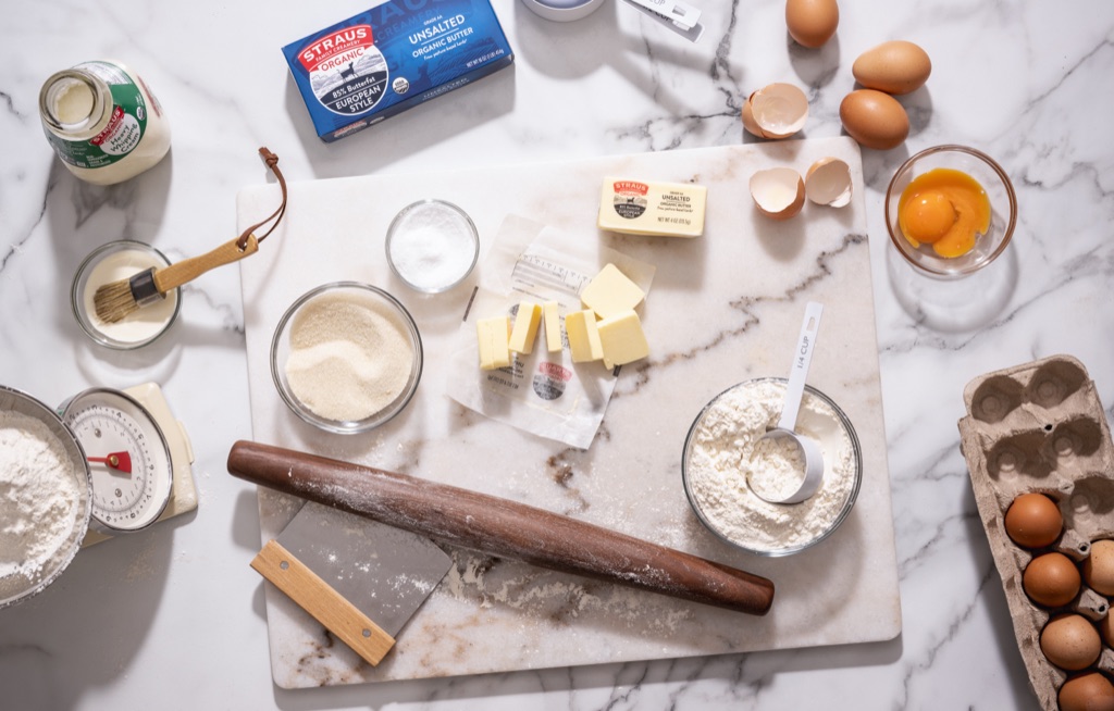 Overhead baking flat lay of Straus European Style butter, eggs, flour, and sugar around a rolling pin on marble