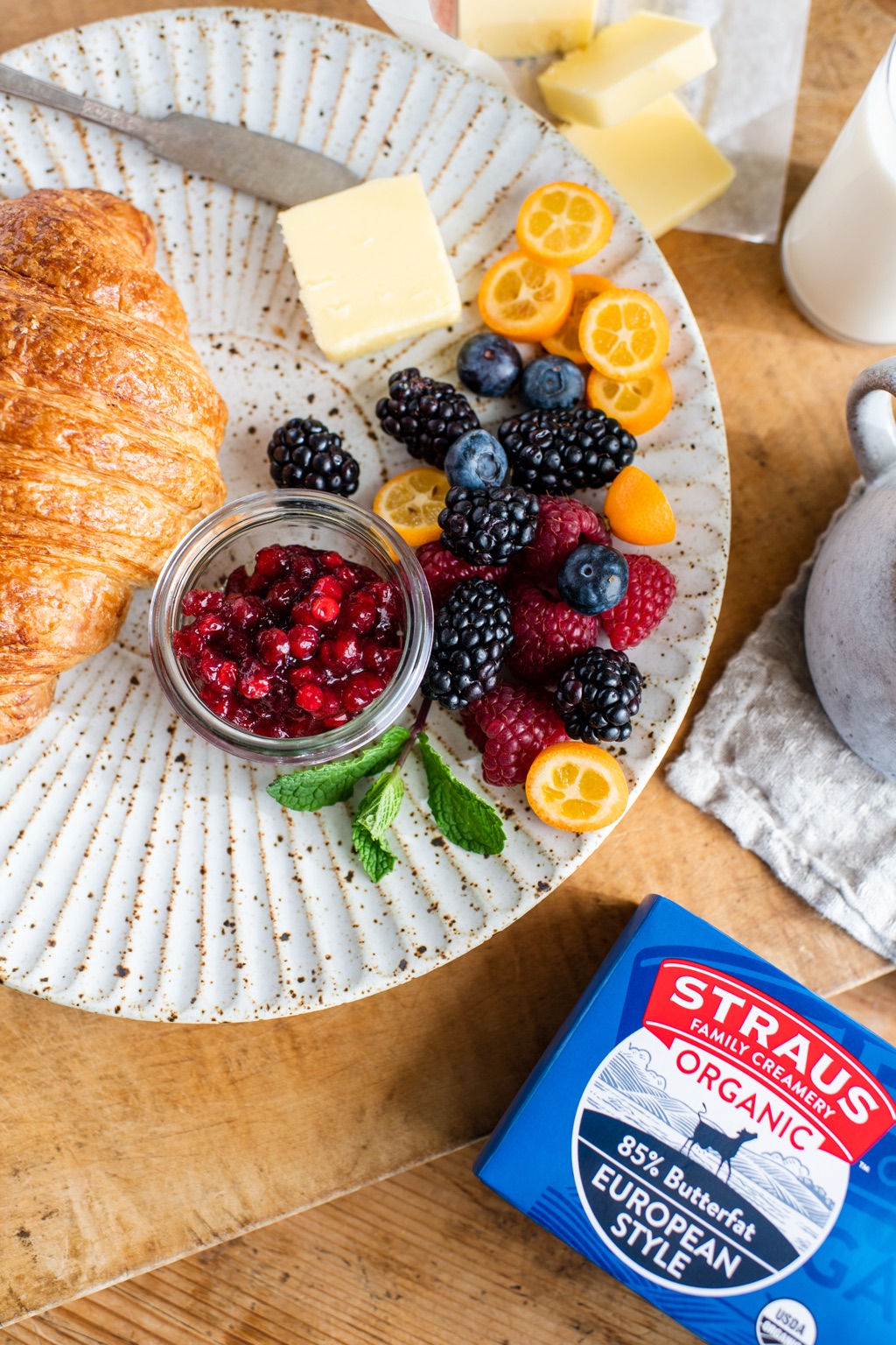 Overhead breakfast plate with a croissant, berries, kumquats, and jam beside a Straus European Style butter box