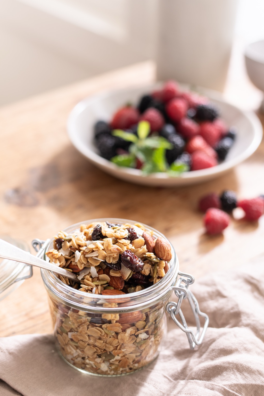 Glass jar of granola with almonds and dried cherries, a bowl of berries behind