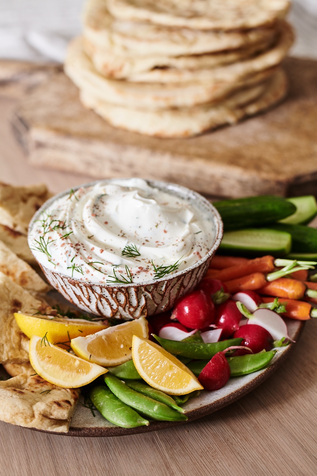 Bowl of creamy yogurt dip with dill surrounded by flatbread, snap peas, radishes, and lemon wedges