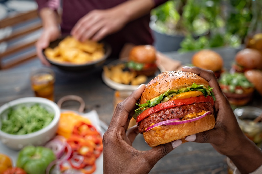 Close-up of hands holding a Sweet Earth Awesome Burger with grilled peppers and arugula at a backyard table