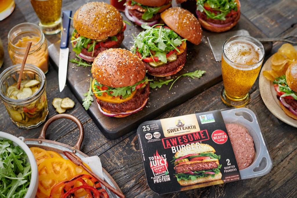 Sweet Earth Awesome Burger package with raw plant-based patties beside a spread of finished burgers, pickles, and beer