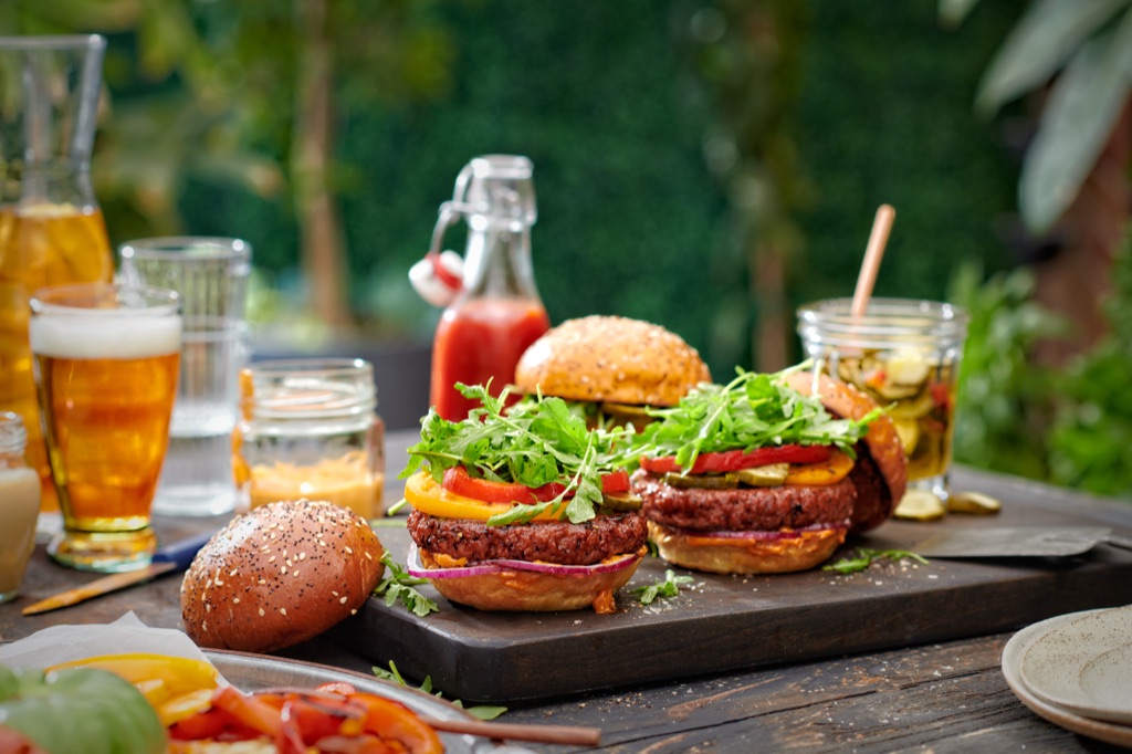 Two Sweet Earth Awesome Burgers piled with arugula and heirloom tomato on a backyard table with beer