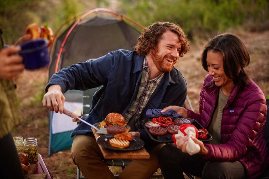 Two friends laughing over a grill tray of plant-based patties and toasted buns at their campsite, photographed by Dawn Heumann