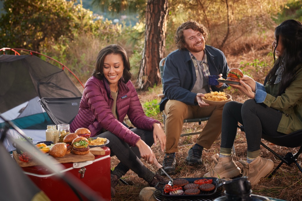 Friends grilling Sweet Earth Awesome Burgers at a campsite, with sliders staged on a red cooler