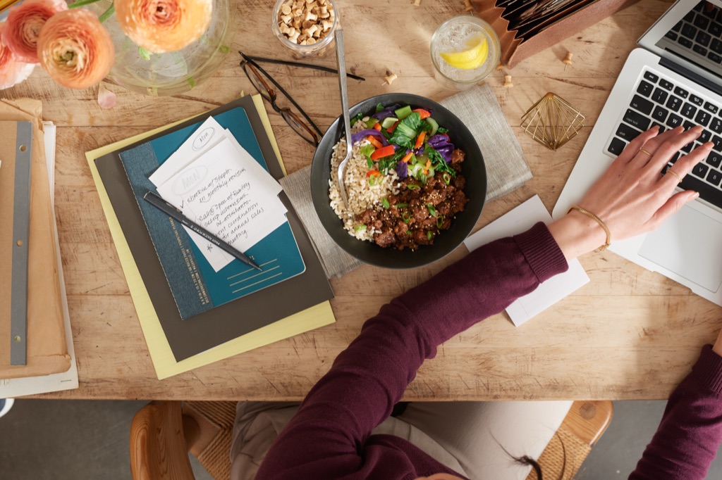 Overhead view of a woman working on a laptop beside a Sweet Earth plant-based ground bowl with brown rice and vegetables