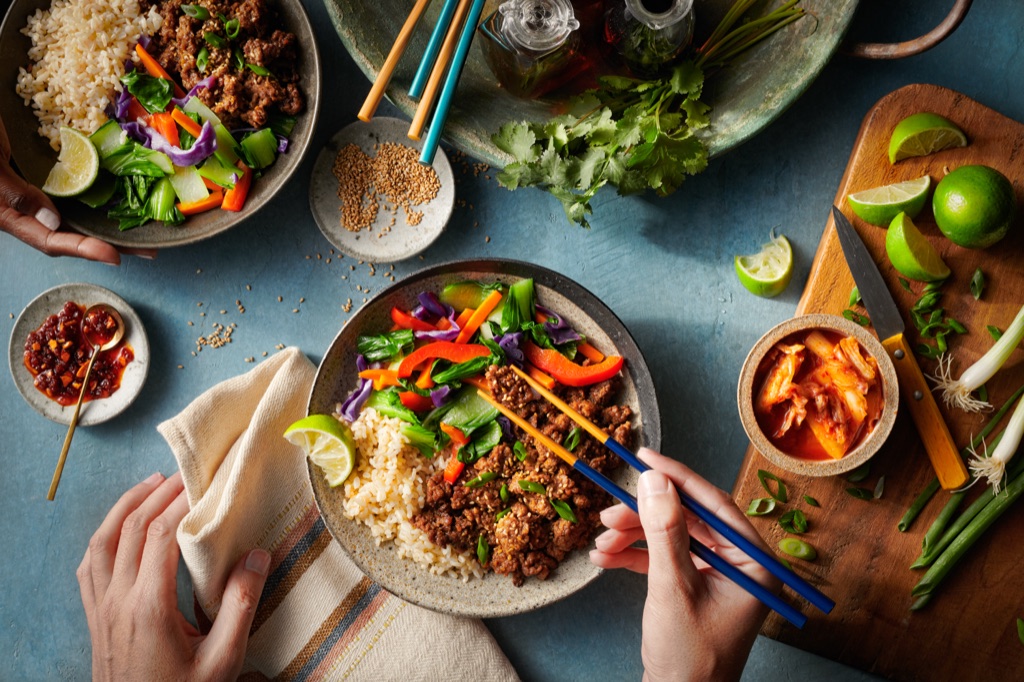 Hands with chopsticks over a bowl of sesame plant-based ground protein, brown rice, and stir-fried vegetables