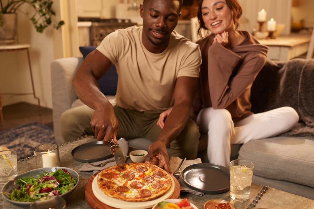 Couple slicing a Sweet Earth veggie pepperoni pizza at a candlelit coffee table, cozy night in