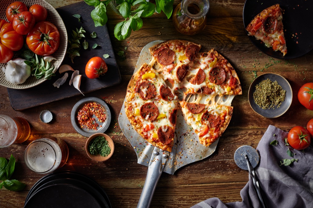Overhead view of a Sweet Earth veggie pepperoni pizza on a metal peel surrounded by tomatoes, basil, and beer