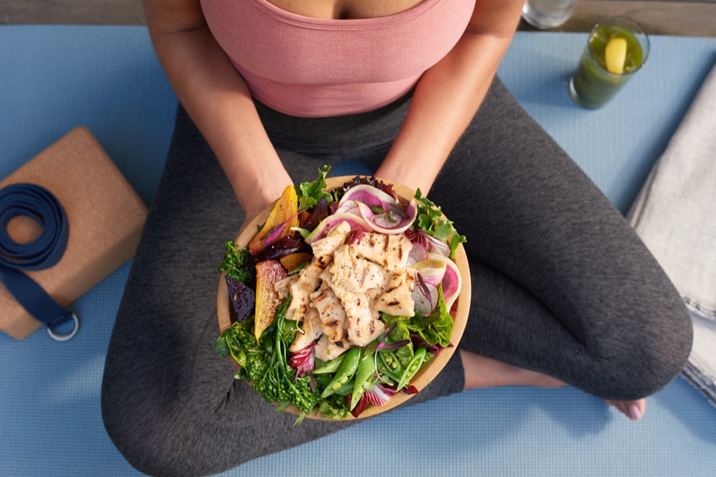 Overhead view of a woman seated cross-legged on a yoga mat holding a chik'n salad bowl with beets and snap peas