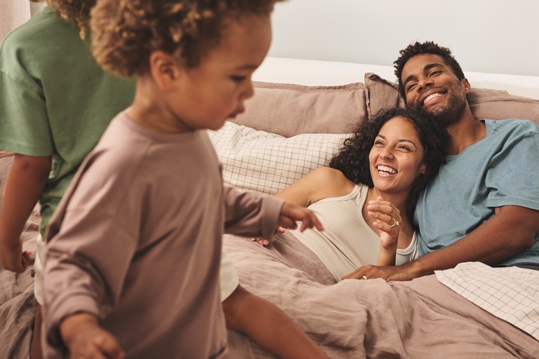 Parents laughing in bed while their kids play in the foreground, Sealy campaign photography, photographed by Dawn Heumann