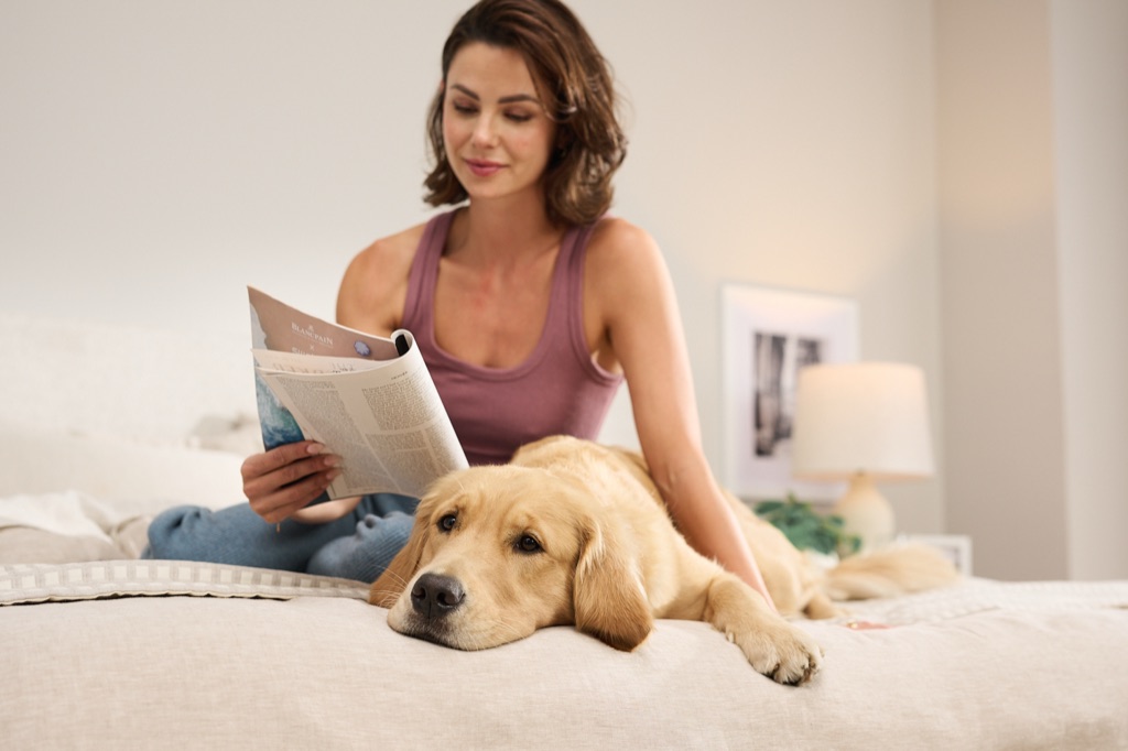 Woman reading a magazine in bed while her golden retriever rests beside her, Sealy lifestyle photography