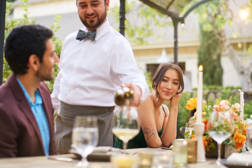 Bow-tied server pouring white wine at a garden dinner party under a wrought iron arbor