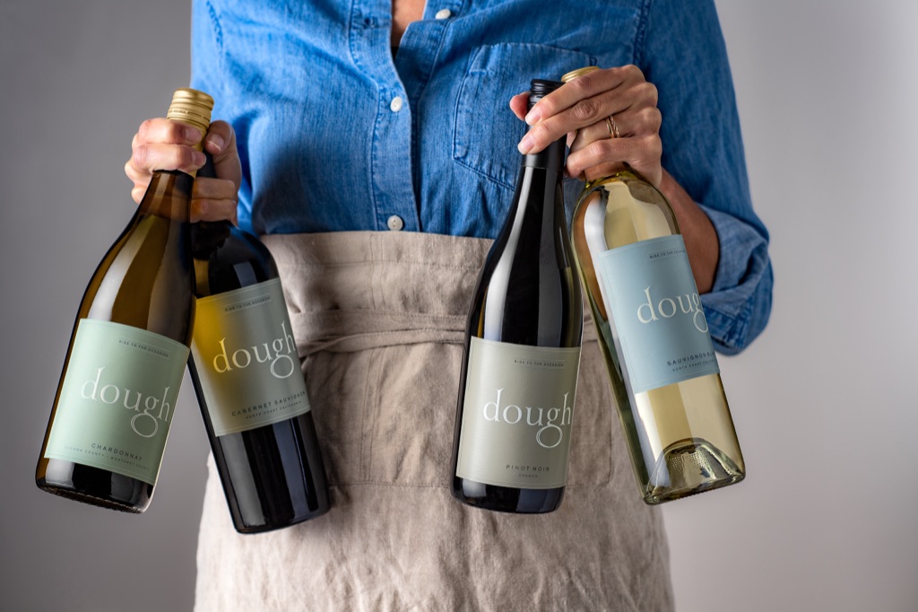 Woman in a linen apron holding four bottles of Dough Wines by the neck