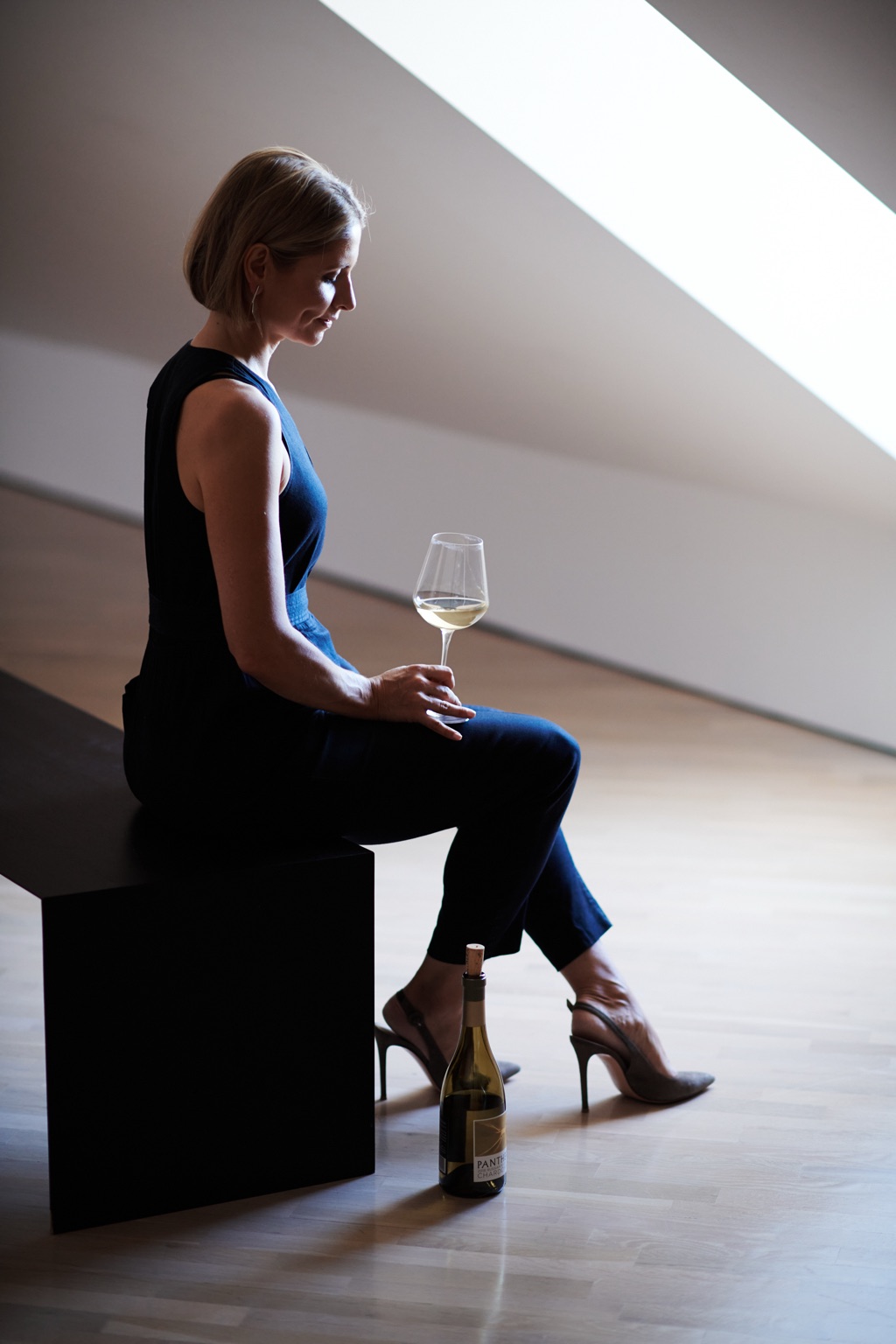 Woman in a navy jumpsuit holding a glass of Panthera chardonnay on a gallery bench under a skylight