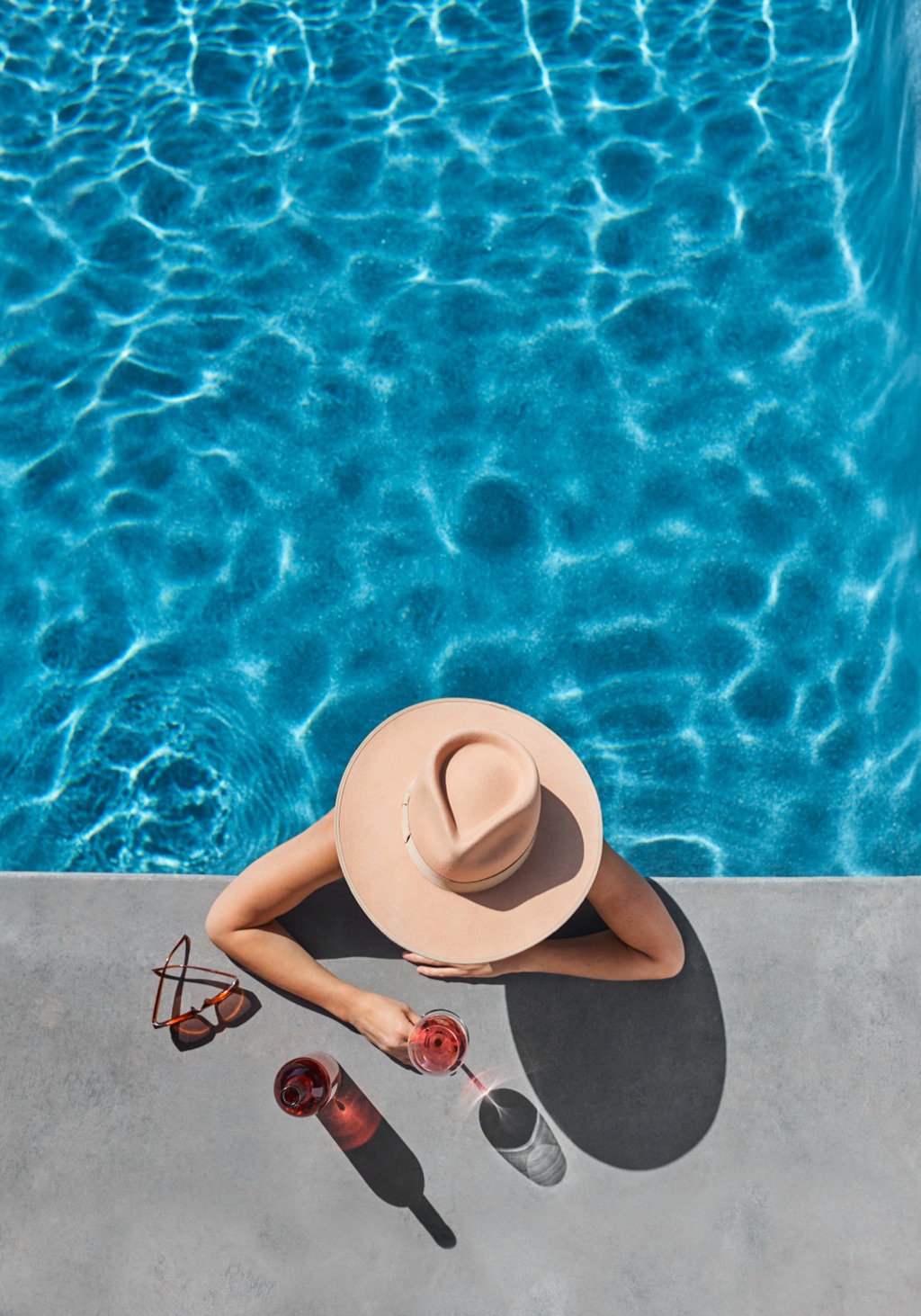 Woman in a sun hat resting at the pool edge with a glass of rose, seen from above