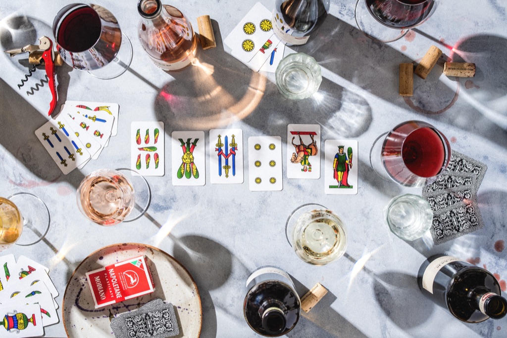 Overhead scopa card game with wine glasses, corks, and afternoon shadows on marble